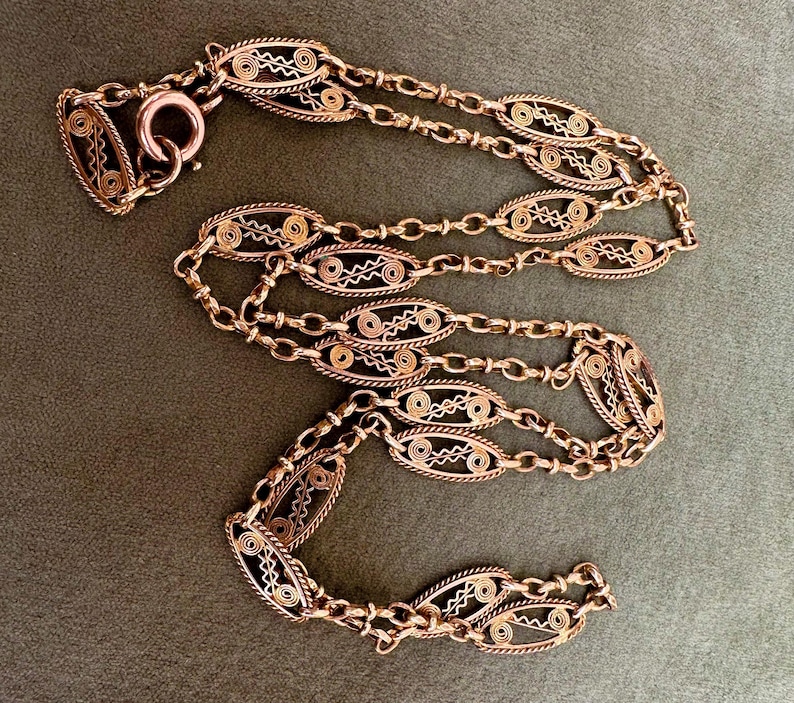 May include: A long, delicate copper chain necklace with intricate oval-shaped links. The chain is made of multiple strands of wire woven together to create a delicate, openwork design.