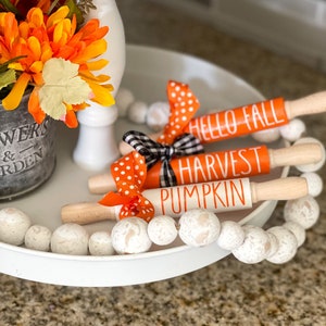 May include: Three wooden rolling pins with orange and white polka dot ribbon bows. The rolling pins are decorated with the words "Hello Fall", "Harvest", and "Pumpkin". The rolling pins are displayed on a white tiered tray with white beads.