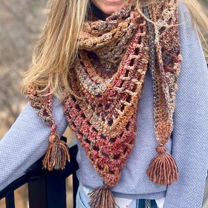 May include: A handmade crochet shawl in shades of brown, red, and orange. The shawl has a triangular shape with a lacy pattern and tassels at the ends. The warm colors and textured design create a cozy and stylish accessory.