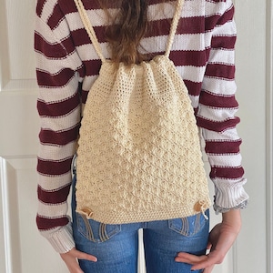 Puede incluir: Una mochila con cordón de ganchillo de color crema con un patrón texturizado. La bolsa tiene correas de color crema y cierres de madera. La persona de la imagen lleva vaqueros azules y un jersey a rayas burdeos y blanco.