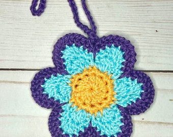 May include: A handmade crochet flower ornament with a loop for hanging. The flower features a vibrant color scheme: a yellow center, surrounded by light blue petals, and outlined with a deep purple border. The ornament is displayed against a light wood background.