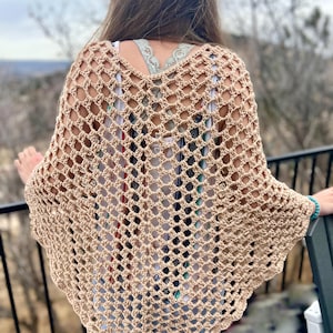 May include: A tan crochet poncho with a diamond-shaped open weave design. The poncho has a scalloped edge and is worn over a white tank top and blue jeans. The person is standing on a balcony overlooking a natural landscape.
