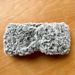 K&ouml;nnte beinhalten: Ein grau-wei&szlig;es Stirnband aus Kunstfell mit einem gedrehten Knotendetail in der Mitte. Das Accessoire besteht aus einem weichen, strukturierten Material mit einem melierten Farbmuster, das eine dichte, flauschige Optik auf der gesamten Oberfl&auml;che erzeugt.