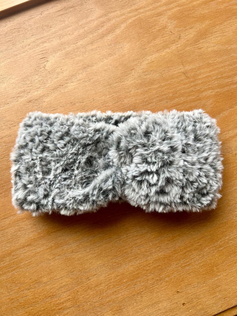 K&ouml;nnte beinhalten: Ein grau-wei&szlig;es Stirnband aus Kunstfell mit einem gedrehten Knotendetail in der Mitte. Das Accessoire besteht aus einem weichen, strukturierten Material mit einem melierten Farbmuster, das eine dichte, flauschige Optik auf der gesamten Oberfl&auml;che erzeugt.