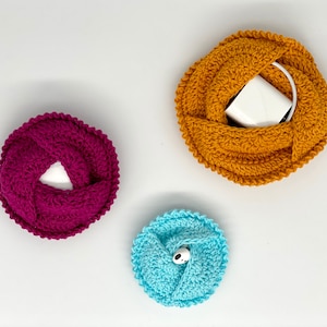 May include: Three handmade crochet cable organizers in shades of magenta, mustard yellow, and light blue. The magenta and mustard yellow organizers are circular, with a cable or charger inside. The light blue organizer is smaller, with a white and black button.