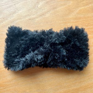 K&ouml;nnte beinhalten: Ein schwarzes Stirnband aus dickem, flauschigem Kunstfellmaterial. Das Accessoire hat eine strukturierte, zottelige Optik mit einem mittigen, gedrehten Knotendetail, das f&uuml;r ein weiches und volumin&ouml;ses Aussehen auf der gesamten Oberfl&auml;che sorgt.