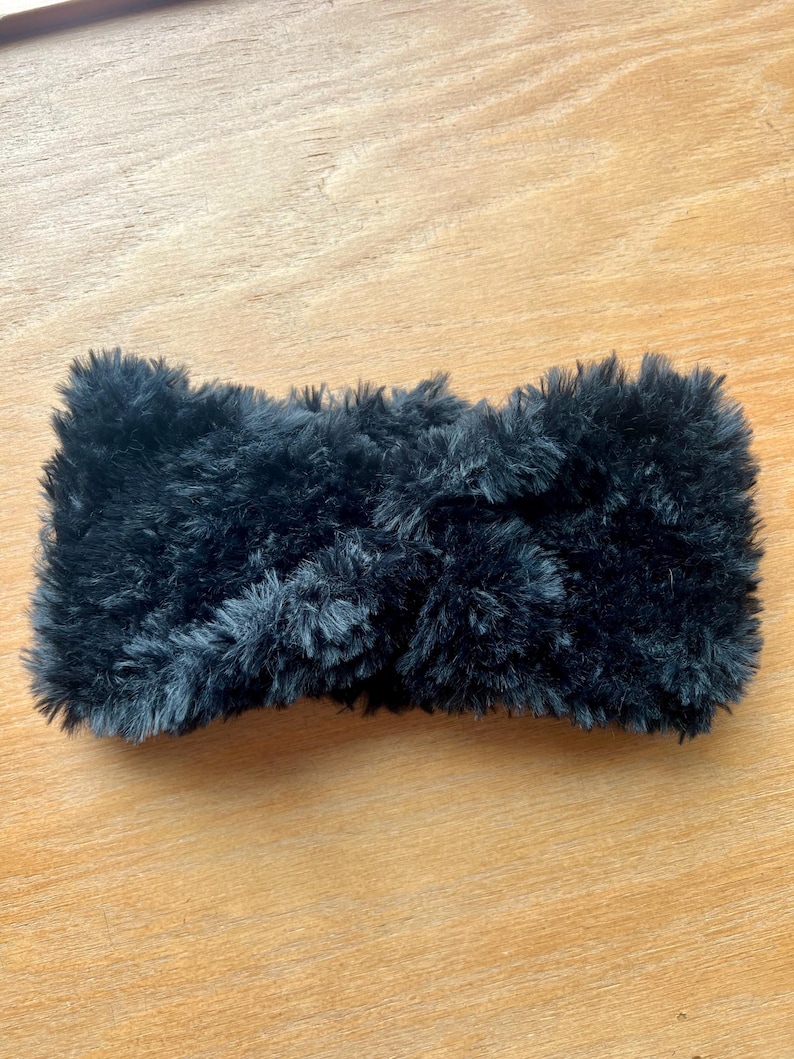 K&ouml;nnte beinhalten: Ein schwarzes Stirnband aus dickem, flauschigem Kunstfellmaterial. Das Accessoire hat eine strukturierte, zottelige Optik mit einem mittigen, gedrehten Knotendetail, das f&uuml;r ein weiches und volumin&ouml;ses Aussehen auf der gesamten Oberfl&auml;che sorgt.