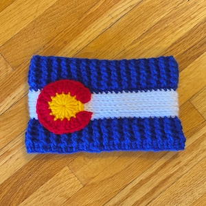 Könnte beinhalten: Gehäkeltes Stirnband in den Farben der Flagge des Bundesstaates Colorado. Das Stirnband ist blau mit einem weißen Streifen und einem roten Kreis mit gelber Mitte.