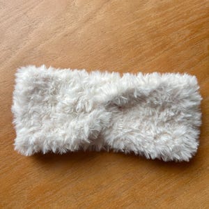 K&ouml;nnte beinhalten: Ein wei&szlig;es Stirnband aus weichem, flauschigem Kunstfellmaterial. Das Accessoire zeichnet sich durch ein markantes, gedrehtes Knotendetail in der Mitte aus, das eine strukturierte und geschichtete Optik auf der gesamten Oberfl&auml;che erzeugt.
