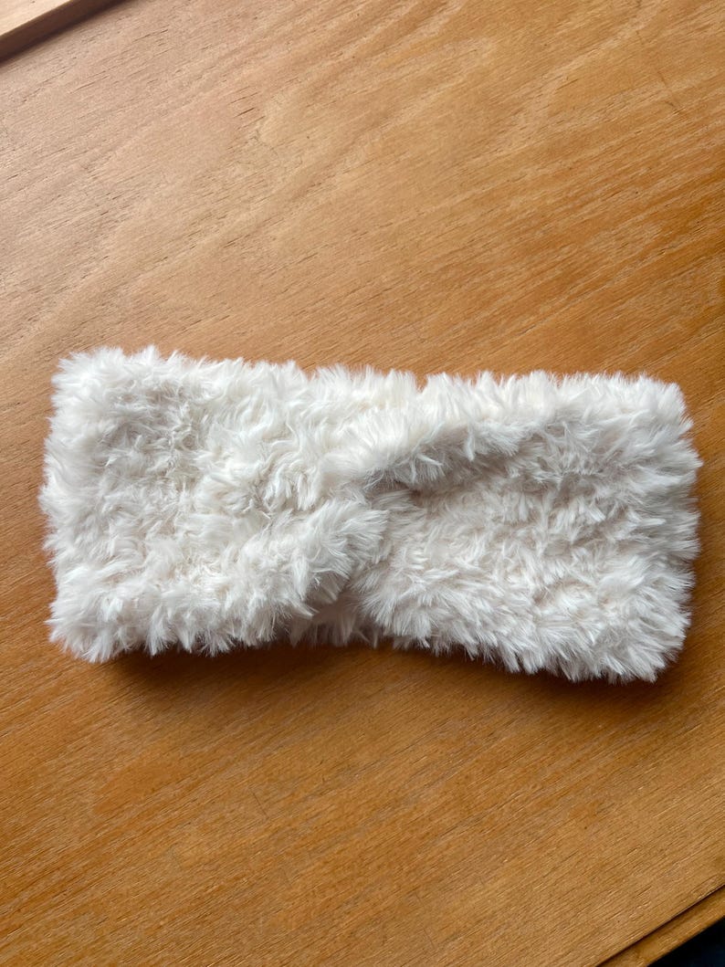K&ouml;nnte beinhalten: Ein wei&szlig;es Stirnband aus weichem, flauschigem Kunstfellmaterial. Das Accessoire zeichnet sich durch ein markantes, gedrehtes Knotendetail in der Mitte aus, das eine strukturierte und geschichtete Optik auf der gesamten Oberfl&auml;che erzeugt.
