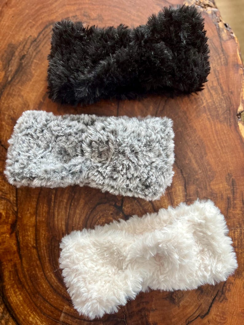 K&ouml;nnte beinhalten: Drei flauschige Stirnb&auml;nder mit einem gedrehten Knoten in der Mitte. Das Set besteht aus einem einfarbigen schwarzen Stirnband, einem grau-wei&szlig; melierten Stirnband und einem cremefarbenen Stirnband aus weichem, pl&uuml;schigem Material.