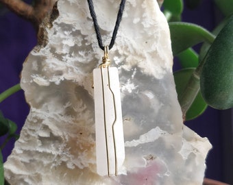May include: A white crystal pendant with gold wire wrapping, hanging from a black cord. The pendant is resting on a piece of white and gray agate.
