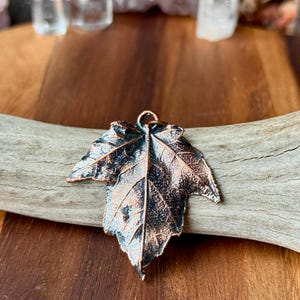 May include: A copper-toned metal pendant shaped like a maple leaf with a dark, oxidized patina in the recessed areas and a bright, metallic finish on the raised veins and edges. The leaf features a small circular loop at the top.