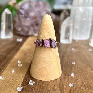 May include: A copper ring featuring three rectangular, raw-cut pink gemstones. The ring is displayed on a wooden cone-shaped stand, with a blurred background of crystals and greenery. The gemstones have a rough, textured appearance.