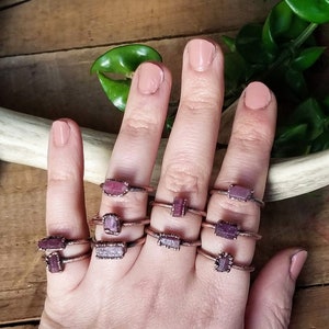 May include: A hand displays several copper-colored rings, each featuring a rectangular, pink gemstone. The rings are set on a simple band. The background includes a wooden surface and green foliage, creating a natural aesthetic.