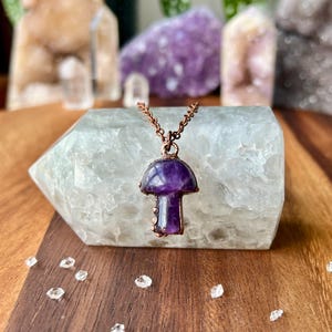 May include: A mushroom-shaped pendant necklace with a purple amethyst gemstone cap and stem, set in a copper-colored metal. The necklace is displayed on a large, light-colored crystal, with other crystals in the background.