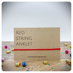 May include: Red string anklet on a brown card with the text "RED STRING ANKLET" and the website address "www.thedreamingbuddha.com".