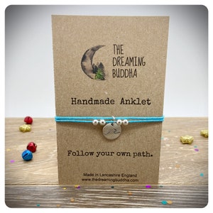 May include: A turquoise blue string anklet with a silver charm featuring a wave design. The anklet is displayed on a brown card with the text "THE DREAMING BUDDHA Handmade Anklet Follow your own path. Made in Lancashire England www.thedreamingbuddha.com"
