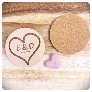 May include: Two round wooden coasters with a heart design and the initials "E & D" and the date "19/06/2014" engraved on one coaster. A small pink felt heart is on the wooden surface.
