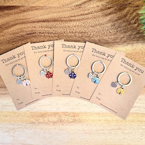 May include: A collection of ladybug keyrings in red, blue, and yellow, presented on kraft paper cards. Each keyring features a silver ring and a heart charm inscribed with "Thank You". The cards are printed with the text "Thank you for helping me grow".