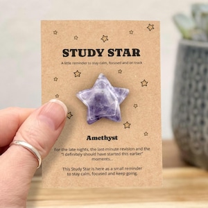 Op de afbeelding: Een paarse amethist ster-vormige kristal op een bruine kaart met de tekst "STUDY STAR". De kaart heeft een boodschap over kalm blijven en gefocust zijn. De ster wordt vastgehouden door een hand met een zilveren ring.