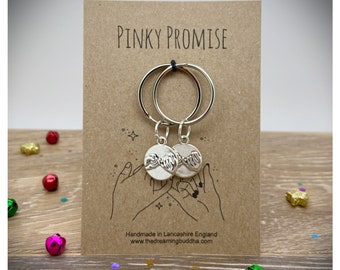 May include: Two silver keychains with interlocking pinky fingers on round charms. The keychains are displayed on a brown card with the text "Pinky Promise" and "Handmade in Lancashire England www.thedreamingbuddha.com".