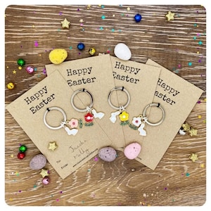 May include: Three brown card tags with the text "Happy Easter" on each. Each tag has a silver key ring with a charm attached. The charms are white bunnies with flowers. The tags are on a wooden surface with colorful confetti.