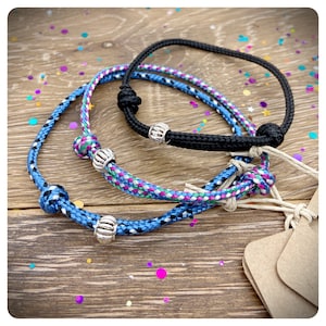 May include: Three rope bracelets in the image. One is black, the other two are blue with colorful accents. Each bracelet has a silver bead. The bracelets are on a wooden surface.