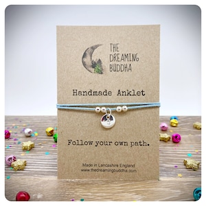 May include: A light blue handmade ankle bracelet with a silver charm featuring the number 8. The bracelet is displayed on a brown card with the text "THE DREAMING BUDDHA Handmade Anklet Follow your own path. Made in Lancashire England www.thedreamingbuddha.com"