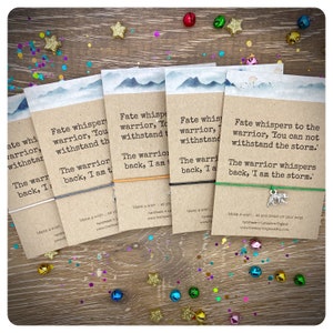 May include: Six colorful wristbands with inspirational messages about being a warrior and facing challenges. The wristbands are made of string and have a charm attached to each one. The messages on the wristbands are: "Fate whispers to the warrior, You can not withstand the storm." and "The warrior whispers back, I am the storm."