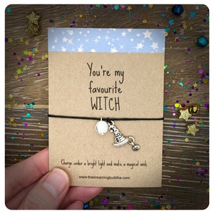 May include: A brown card with the text "You're my favourite WITCH" and a black string with a white bead and a silver charm of a witch's hat. The text below the card reads "Charge under a bright light and make a magical wish. www.thedreamingbuddha.com"