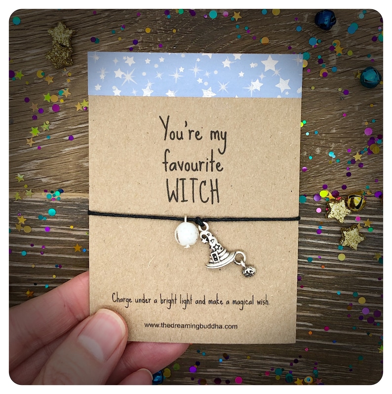 May include: A brown card with the text "You're my favourite WITCH" and a black string with a white bead and a silver charm of a witch's hat. The text below the card reads "Charge under a bright light and make a magical wish. www.thedreamingbuddha.com"