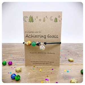 Könnte beinhalten: Eine Karte mit dem Text "A crystal wish for Achieving Goals" und ein Armband mit grünen, braunen und schwarzen Perlen und einem silbernen Anhänger. Die Karte zeigt eine Zeichnung von Bäumen und Sternen. Das Armband ist handgefertigt in Lancashire, England.