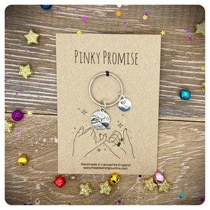 May include: A silver keychain with two charms. One charm is a circle with two hands making a pinky promise. The other charm is a smaller circle with a design. The keychain is on a brown card with the text "Pinky Promise" and "Handmade in Lancashire England www.thedreamingbuddha.com".