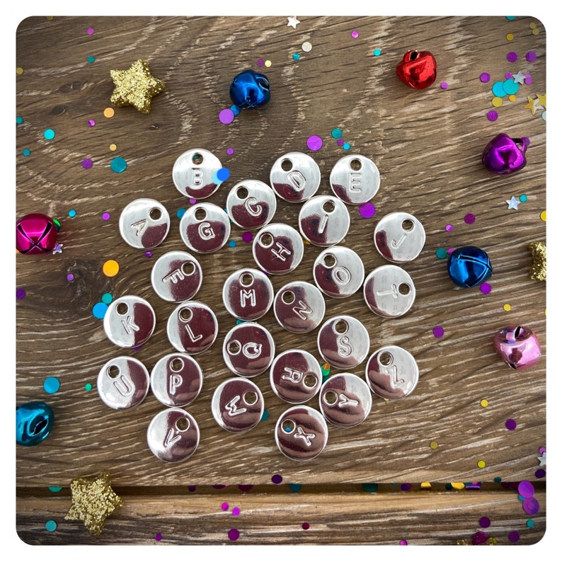 May include: A collection of silver metal letter charms with a variety of letters of the alphabet. The charms are arranged on a wooden surface with colorful confetti.
