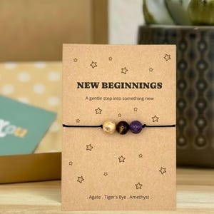 Puede incluir: Pulsera con tres cuentas de piedras preciosas: ágata, ojo de tigre y amatista, ensartadas en un cordón negro. Se muestra en una tarjeta con el texto "NEW BEGINNINGS" e ilustraciones de estrellas. Las cuentas miden aprox. 0,8 cm.