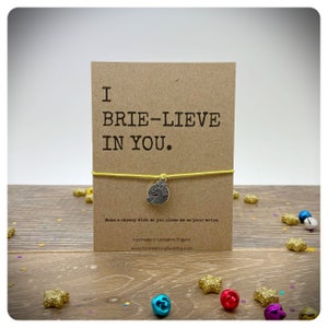 Puede incluir: Una pulsera de amistad amarilla con un dije de plata que dice "cheese". La pulsera está unida a una tarjeta que dice "I brie-lieve in you."