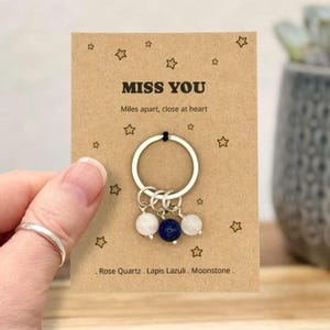 Könnte beinhalten: Ein silberner Schlüsselanhänger mit drei Edelsteinanhängern: Rosenquarz, Lapislazuli und Mondstein. Der Schlüsselanhänger ist an einer braunen Karte mit dem Text "MISS YOU" und "Miles apart, close at heart" befestigt.