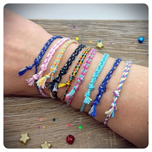 May include: A set of 10 colourful braided friendship bracelets with tassels. The bracelets are made of thin, colourful threads and are tied in a knot at the wrist. The bracelets are displayed on a person's wrist.
