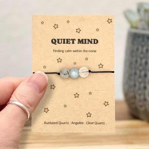 May include: A bracelet featuring three gemstone beads: rutilated quartz, angelite, and clear quartz. The beads are strung on a black cord and presented on a card. The card reads "QUIET MIND" and "Finding calm within the noise", with star motifs.