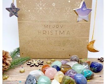 Op de afbeelding: Een bruine kartonnen doos met een kerst-adventskalenderontwerp. De doos heeft een gouden folie-inscriptie met de tekst "Merry Christmas". De doos is omringd door verschillende gekleurde kiezels.