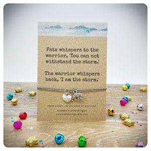 May include: A gray string bracelet with a silver charm that says "B" and a silver bear charm. The bracelet is on a card with the text "Fate whispers to the warrior, 'You can not withstand the storm.' The warrior whispers back, 'I am the storm.' Make a wish... as you place on your wrist. handmade in Lancashire England www.thedreamingbuddha.com"
