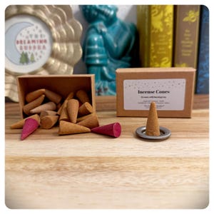 May include: A box of incense cones with a brown paper label that reads "Incense Cones 20 cones with burning tray". The box is sitting on a wooden surface with a few incense cones scattered around it. One incense cone is sitting on a silver metal tray.