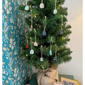May include: A small artificial Christmas tree decorated with various coloured gemstone ornaments. The tree is in a burlap sack and is sitting on a table with a lit candle and a box of small candles.