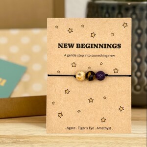 May include: A bracelet featuring three beads—a beige agate, a brown tiger's eye, and a purple amethyst—on a black cord, displayed on a tan card printed with stars, the text "NEW BEGINNINGS", "A gentle step into something new", and "Agate . Tiger's Eye . Amethyst".
