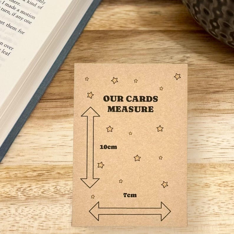 May include: A rectangular kraft paper card with the text "OUR CARDS MEASURE". The card dimensions are indicated with arrows, showing 10cm in height and 7cm in width. Small star illustrations surround the text and measurements.