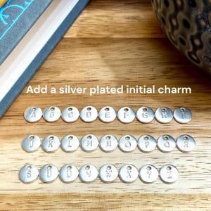 May include: Silver-plated initial charms, each with a different letter from A to Z. The charms are arranged in three rows on a wooden surface. The text "Add a silver plated initial charm" is above the charms.