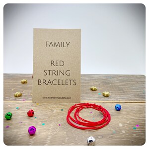 May include: Red string bracelets with the words "Family Red String Bracelets" on a brown card. The card also includes the website address "www.thedreamingbuddha.com".