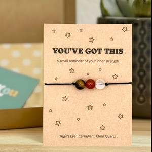 May include: A beaded bracelet with Tiger's Eye, Carnelian, and Clear Quartz stones on a black cord, displayed on a card with the text "YOU'VE GOT THIS". The card has star illustrations and the text "A small reminder of your inner strength".