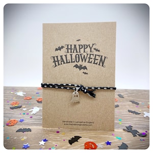 May include: A Halloween bracelet featuring a black and white cord and a silver ghost charm. The bracelet is presented on a brown card printed with "HAPPY HALLOWEEN" and bat motifs. Handmade in Lancashire, England.